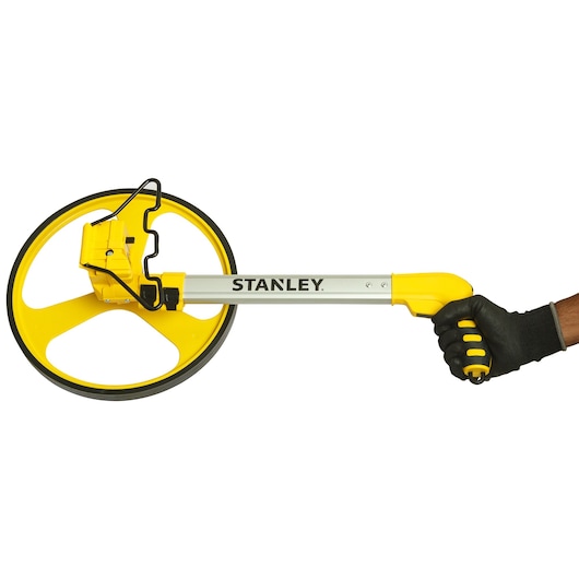 STANLEY 1-77-174 measuring wheel, yellow and black, side angle, held by gloved hand.