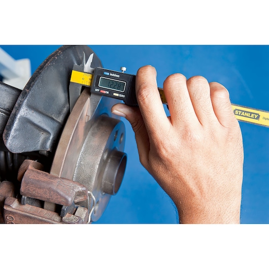 Photo of STANLEY 78-440LA digital caliper measuring a brake disc, close-up on hand and tool, blue background.