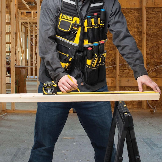 Photo of STANLEY FMST530201 tool vest with multiple pockets holding tools, worn by a person marking wood in a workshop.