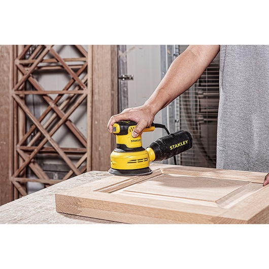 STANLEY SS30-B2C random orbital sander shown sanding a wooden cabinet door in a workshop.