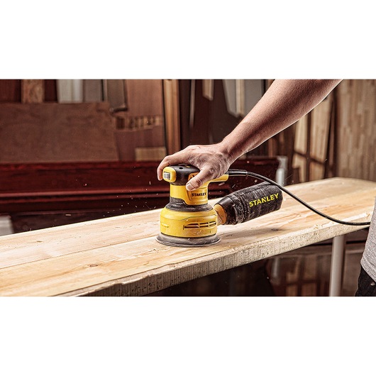 Photo of STANLEY SS30-B2C random orbital sander in use on wooden plank, handheld, dust bag attached.
