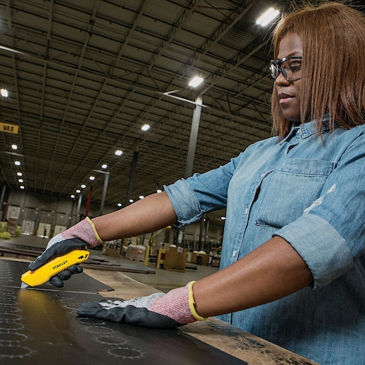 Photo of person in denim shirt using STANLEY STHT10368 utility knife to cut material on work table in warehouse.