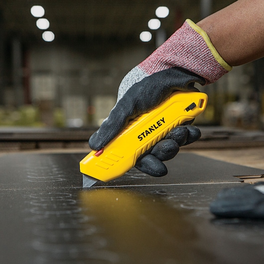 Photo of STANLEY STHT10368 utility knife cutting material, held by a gloved hand in an industrial setting.