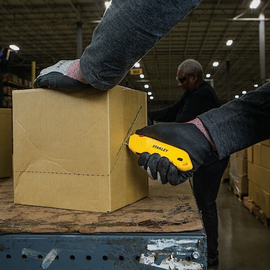Stanley STHT10368 utility knife cutting cardboard box, warehouse setting, worker wearing black gloves.