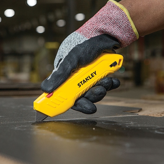 Photo of STANLEY STHT10368 yellow utility knife held by gloved hand cutting material on workbench.