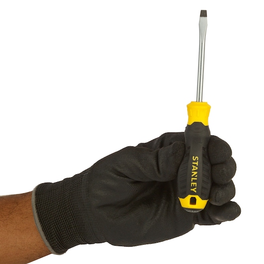A person wearing a black work glove holds a Stanley brand flathead screwdriver with a yellow and black handle, against a white background.